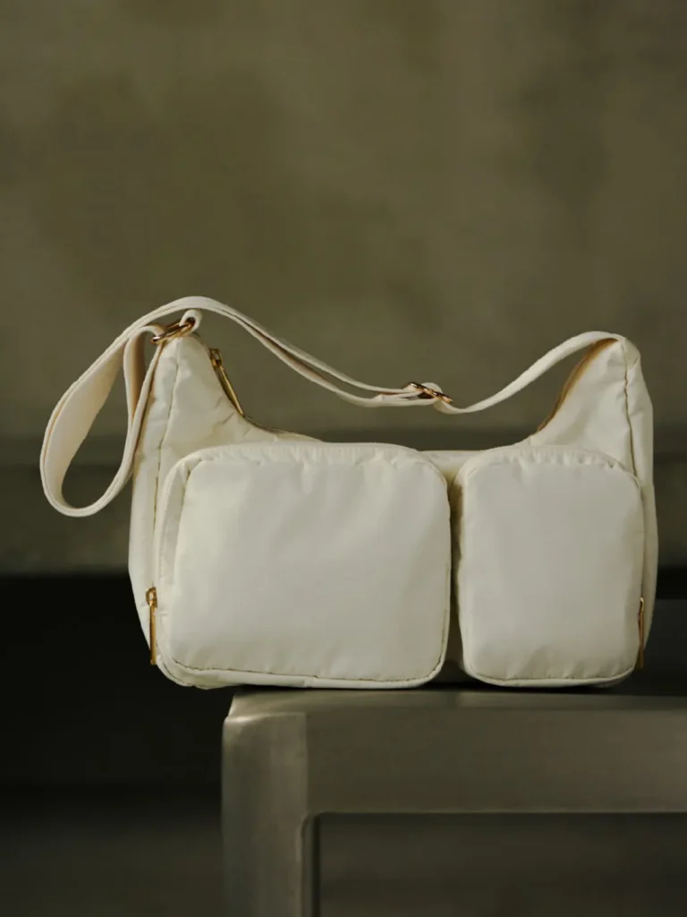 White LeSportsac shoulder bag with oversized padded front pockets displayed on a pedestal, highlighting its soft quilted construction and gold zipper accent