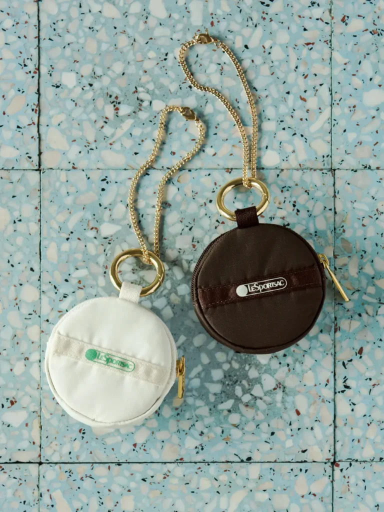 White and brown circular LeSportsac zip pouches with gold chain attachments displayed on a terrazzo surface beside wireless earbud