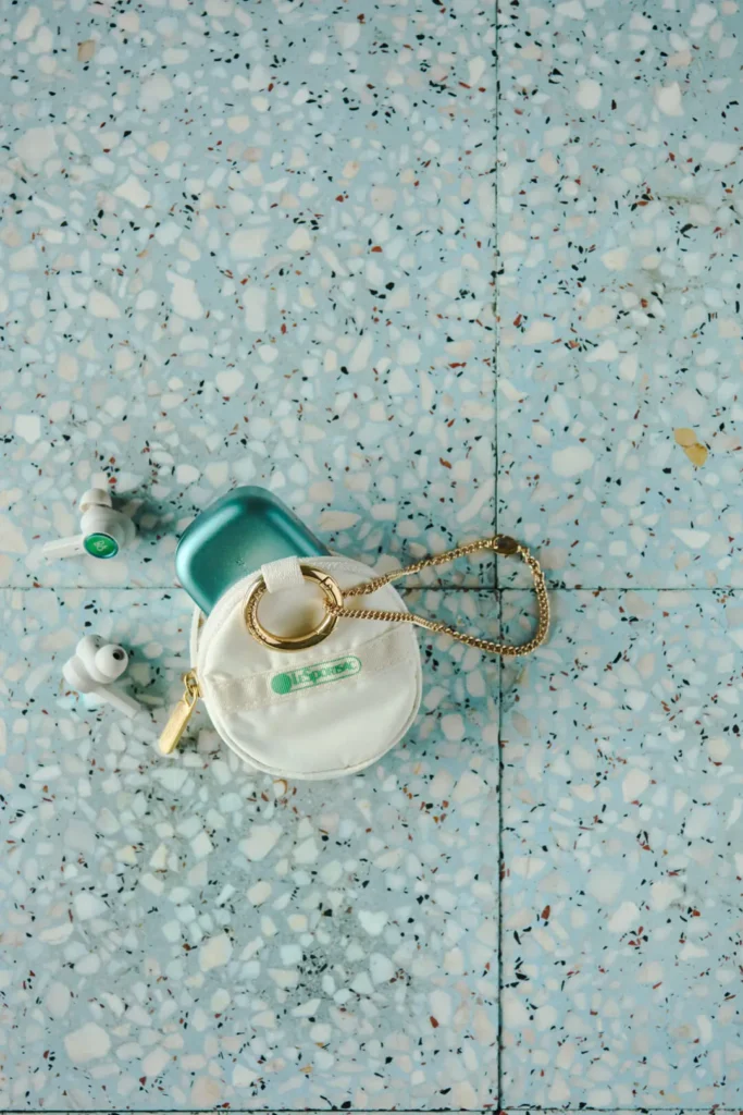 White circular LeSportsac zip pouch holding a teal wireless earbud charging case, arranged on a terrazzo floor with earbuds nearby