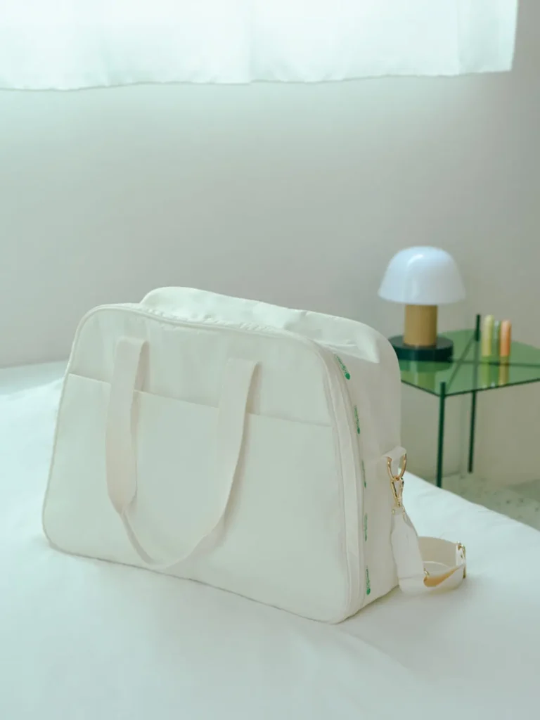 White LeSportsac travel duffel displayed on a neatly made bed in a minimalist room with a glass side table and modern lamp