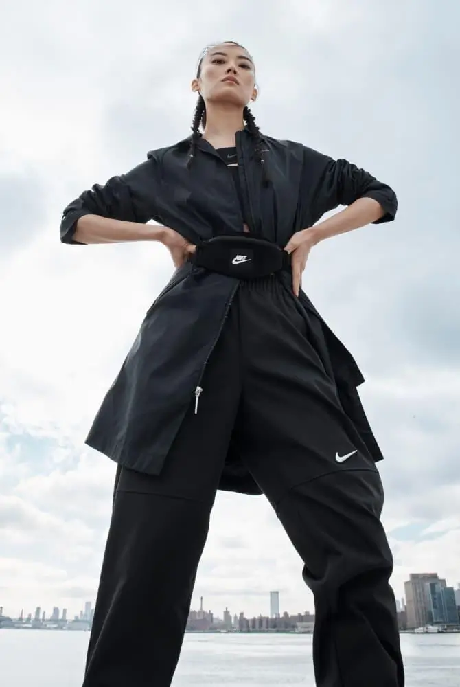 Model wearing an all-black Nike outfit featuring a lightweight longline zip jacket, wide-leg performance pants, and a matching Nike waist bag, photographed from a low angle against a cloudy waterfront skyline.