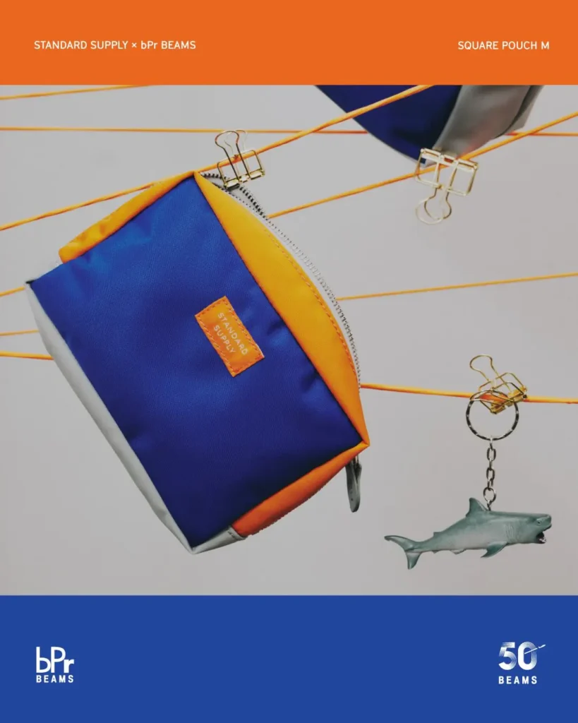STANDARD SUPPLY x bPr BEAMS Square Pouch M in blue, orange, and silver ballistic-style fabric with a zip closure and woven logo patch, displayed against a playful suspended backdrop