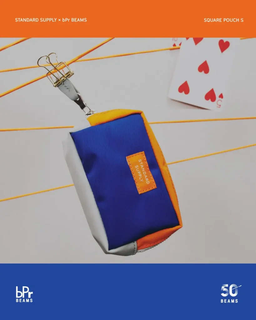 STANDARD SUPPLY x bPr BEAMS Square Pouch S in blue, orange, and silver with a compact zip design and logo patch, suspended among cords and whimsical display props for the BEAMS 50th anniversary collection