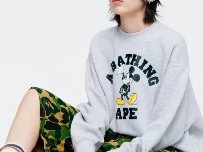 A model wears a light grey A Bathing Ape crewneck sweatshirt featuring Mickey Mouse in camouflage, paired with classic BAPE camo shorts, striped socks, sneakers, and a branded baseball cap in a clean studio set
