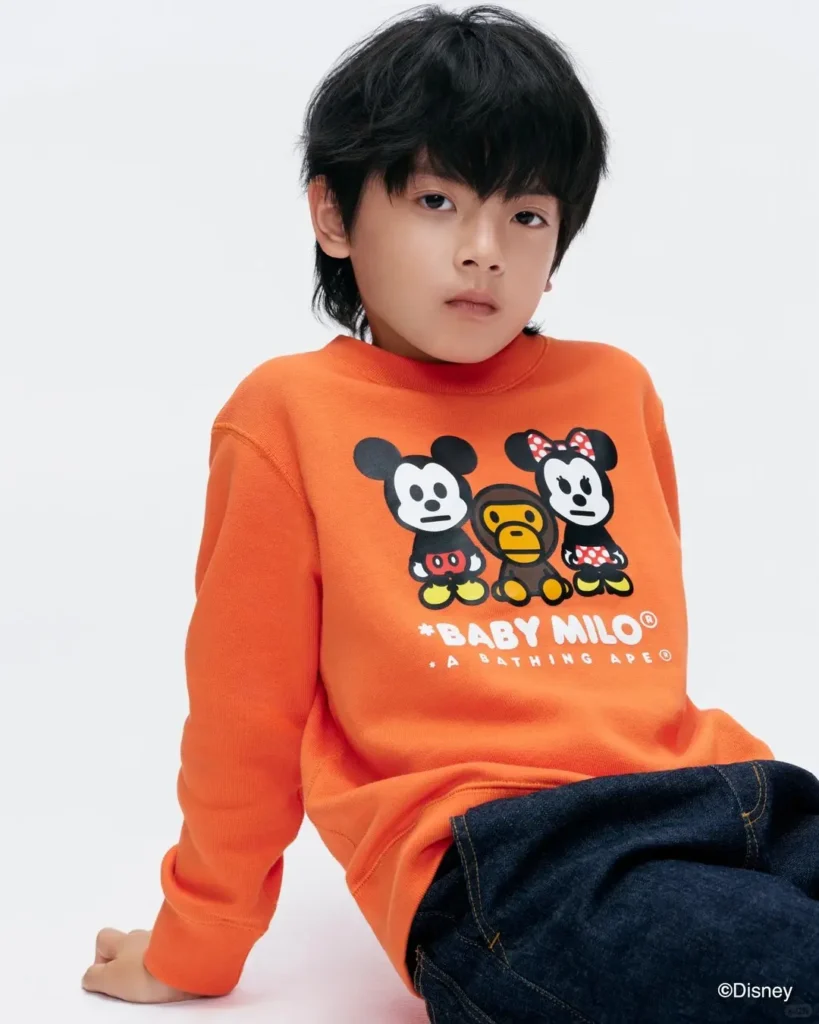 A child models a bright orange Baby Milo sweatshirt featuring stylized Mickey Mouse, Minnie Mouse, and Baby Milo graphics, paired with dark denim jeans against a neutral white studio backdrop