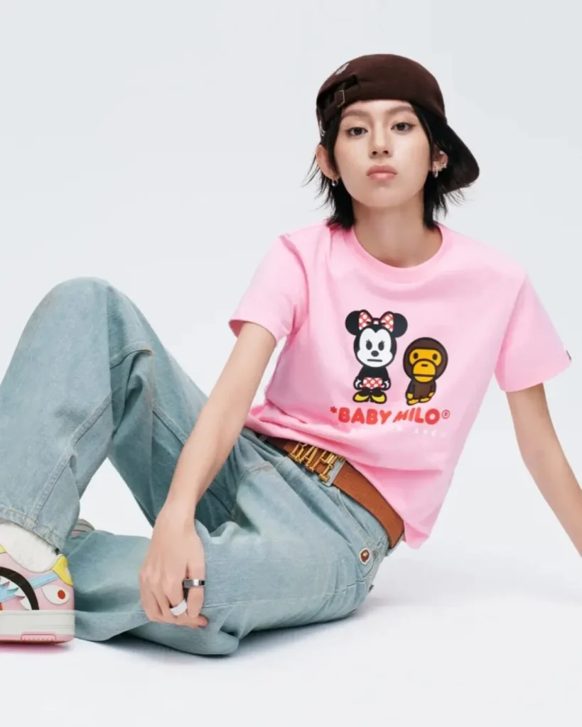 A model wears a pastel pink Baby Milo T-shirt featuring Minnie Mouse alongside Baby Milo, styled with light-wash jeans, a brown belt, and a brown baseball cap in a minimalist studio portrait