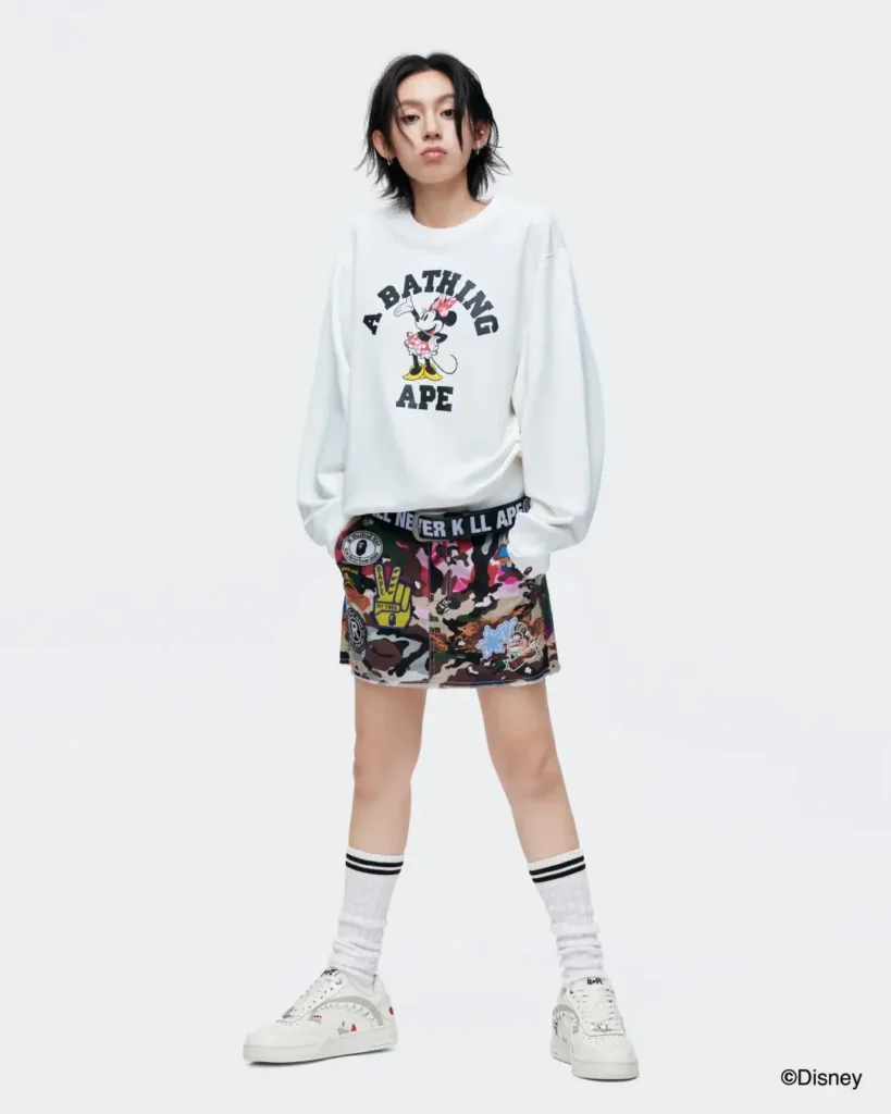 A full-body look showcases a white A Bathing Ape Minnie Mouse sweatshirt paired with a camouflage patchwork mini skirt, striped crew socks, and white BAPE shoe in a studio environment