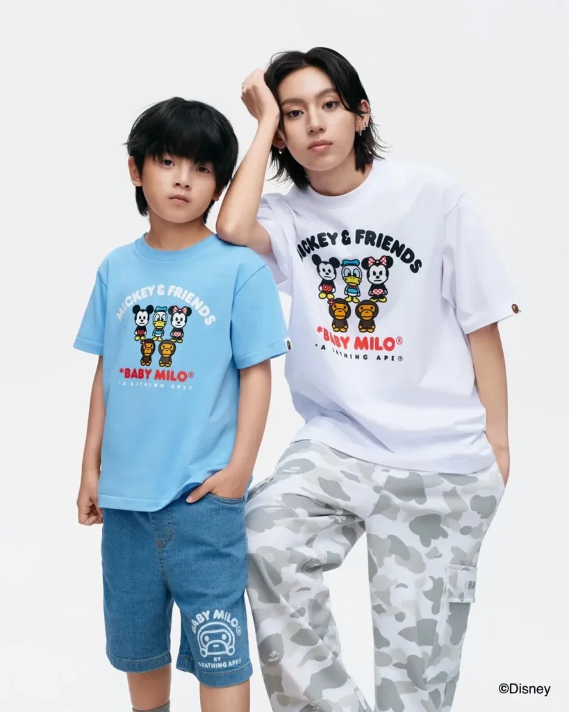 An adult and child stand side by side wearing matching Disney x Baby Milo graphic T-shirts featuring Mickey, Donald Duck, Minnie Mouse, and Baby Milo, styled with denim shorts and grey camouflage cargo pant