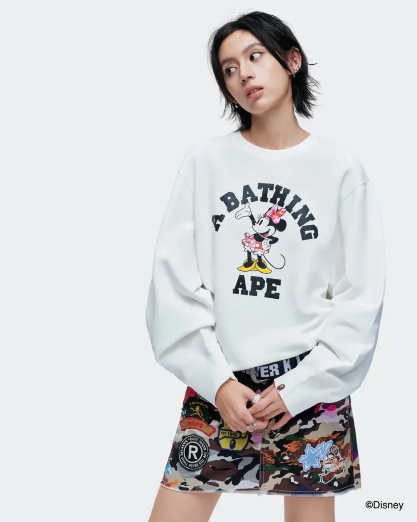 A model wears an oversized white A Bathing Ape sweatshirt with a large Minnie Mouse graphic, paired with a colorful camouflage patchwork skirt decorated with BAPE-inspired patches and badge