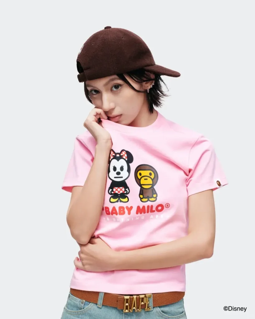 A close-up portrait shows a model wearing a pastel pink Baby Milo and Minnie Mouse graphic T-shirt, styled with light blue jeans, a brown leather belt, multiple rings, earrings, and a sideways brown baseball cap against a clean studio backgroun