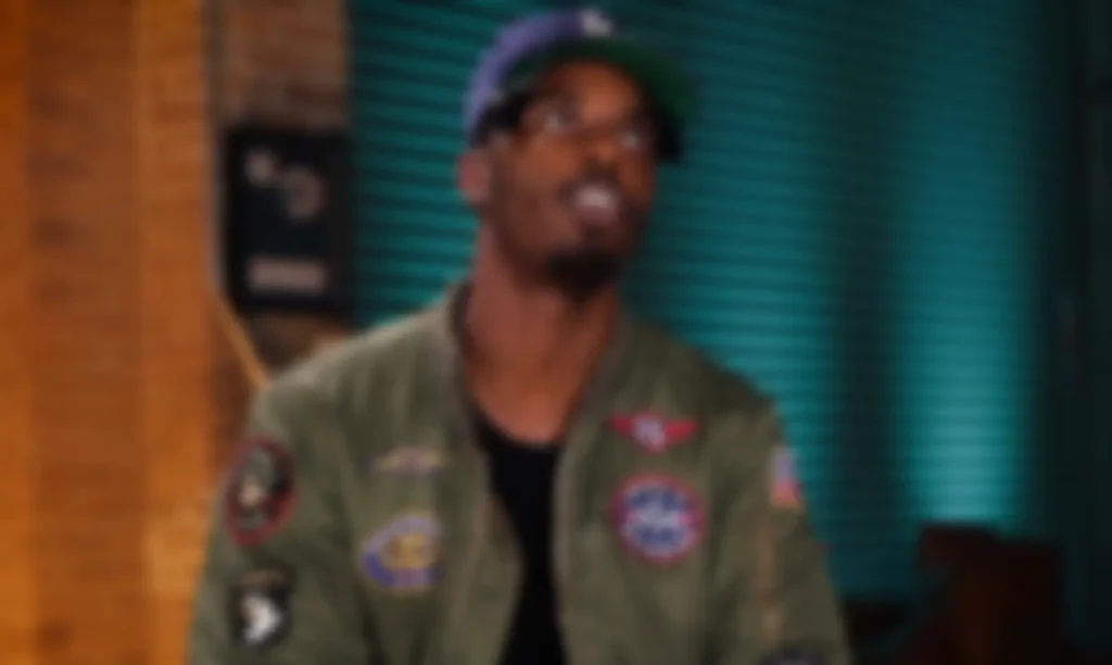 Heavily blurred image of Sean Leon wearing a dark baseball cap and an olive green bomber jacket with embroidered patches, seated indoors against a teal slatted wall and exposed brick background. The intentionally soft focus obscures facial features and fine details while preserving the overall interview-style composition