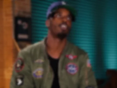 Heavily blurred image of Sean Leon wearing a dark baseball cap and an olive green bomber jacket with embroidered patches, seated indoors against a teal slatted wall and exposed brick background. The intentionally soft focus obscures facial features and fine details while preserving the overall interview-style composition