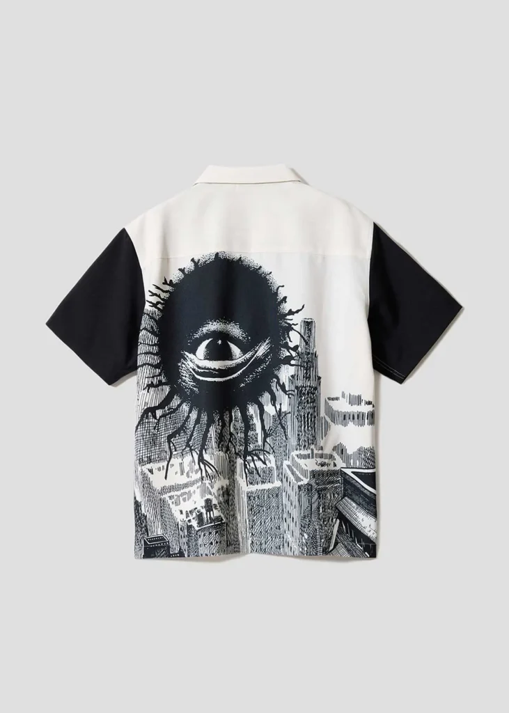 White-and-black short-sleeve shirt featuring a dramatic oversized black eye graphic hovering above a monochrome cityscape across the back