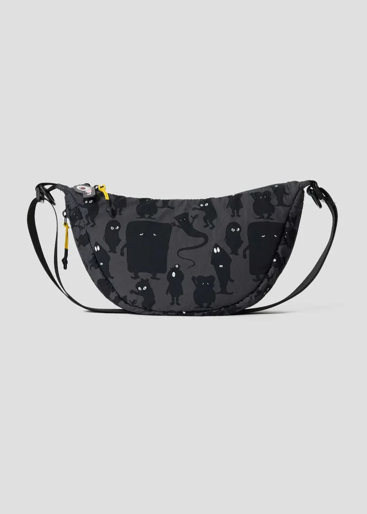 Crescent-shaped black shoulder bag covered in tonal silhouettes of GeGeGe no Kitarō yokai with subtle eye details and a decorative Eyeball Father zipper charm