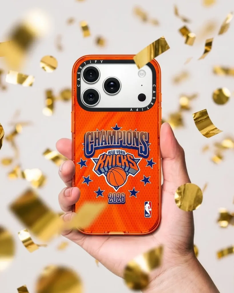 A hand holds up an orange Casetify iPhone case featuring a "Champions New York Knicks 2026" graphic with navy stars and the NBA logo, surrounded by falling gold confetti