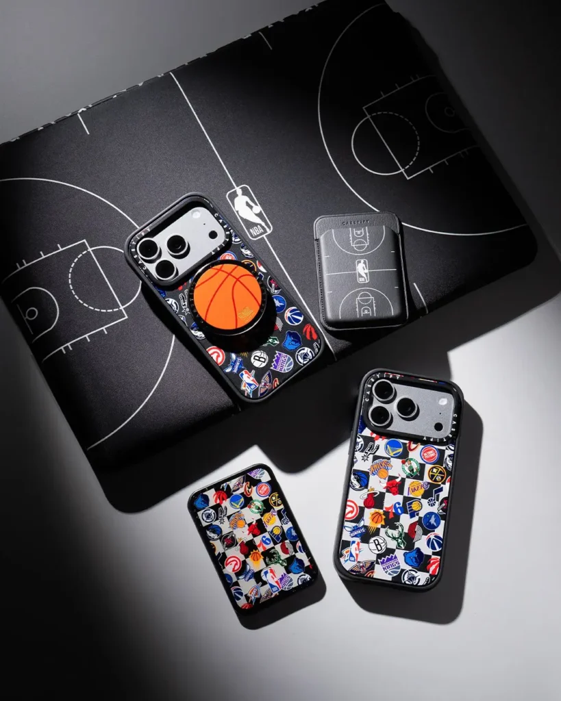 Overhead flat-lay on a black basketball court-print backdrop showing an iPhone case with a team-logo checkerboard pattern and a basketball-shaped MagSafe grip, a black NBA court-print card holder, and two additional phone cases in the same multi-team checkerboard design