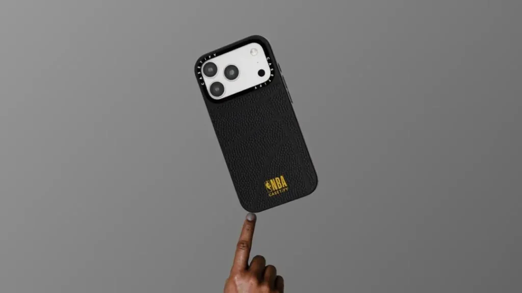 Close-up of a black iPhone case with a textured basketball-leather pattern and a gold "NBA Casetify" logo near the bottom, held up by a fingertip against a gray background