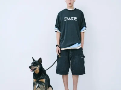 Full-body lookbook image of a model wearing a charcoal ENNOY oversized T-shirt, washed black carpenter shorts, black cap, flip-flops, and layered shirts while standing beside a leashed black-and-tan dog against a white studio backdrop