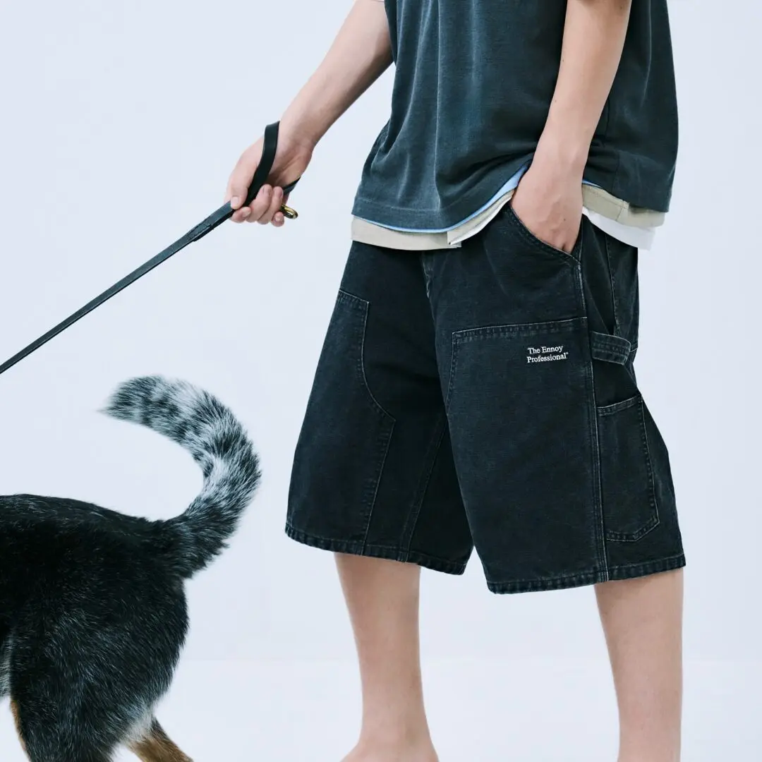 Close-up of washed black ENNOY carpenter shorts with oversized utility pockets and embroidered branding, styled with a layered charcoal T-shirt while the model holds a dog leash.