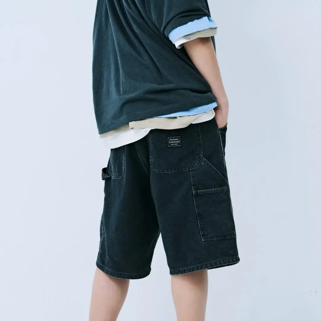 Rear view of the washed black carpenter shorts highlighting oversized back pockets, woven brand patch, utility pocket construction, and faded denim finish beneath layered oversized top