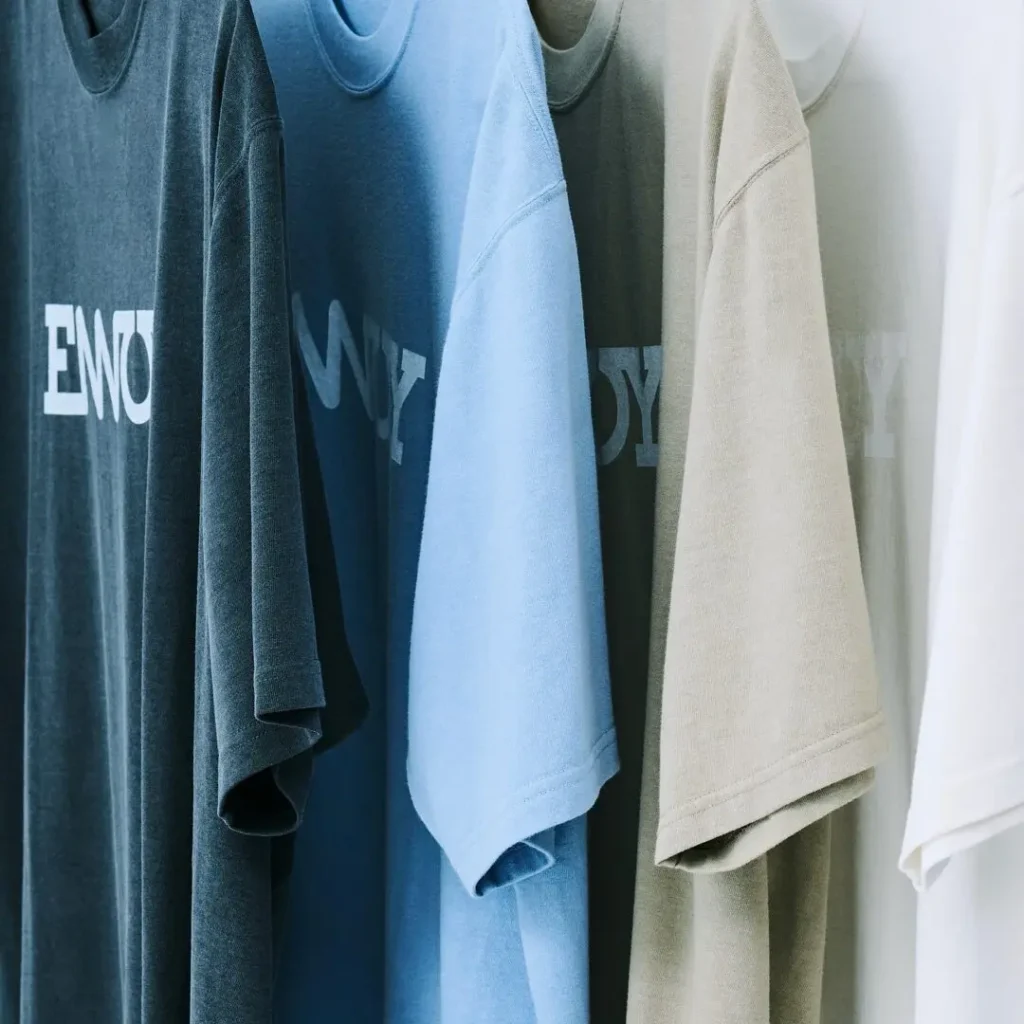 A rack displaying oversized ENNOY logo Tee in washed charcoal, sky blue, olive, beige, and white colorways, emphasizing the heavyweight cotton fabric and relaxed sil
