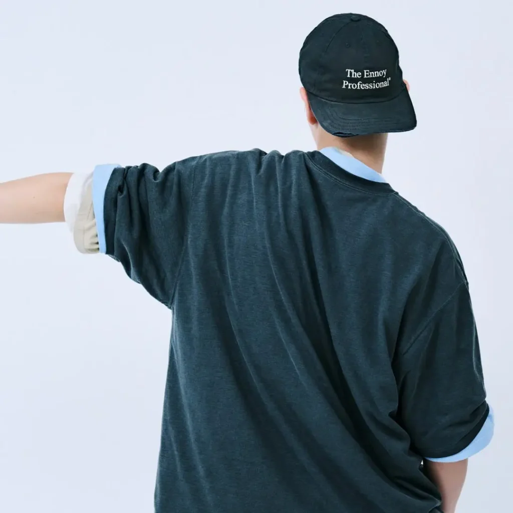 Rear view of a model wear the oversized charcoal ENNOY T-shirt and black embroidered "The Ennoy Professional" cap, showing the relaxed drape, rolled sleeves, and layered styling against a clean studio background