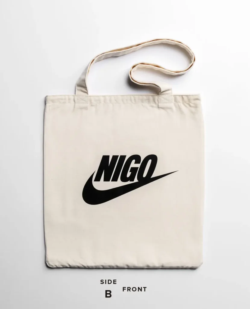 Natural cotton canvas tote bag featuring a bold black NIGO logo inspired by Nike branding centered on the front with long shoulder strap