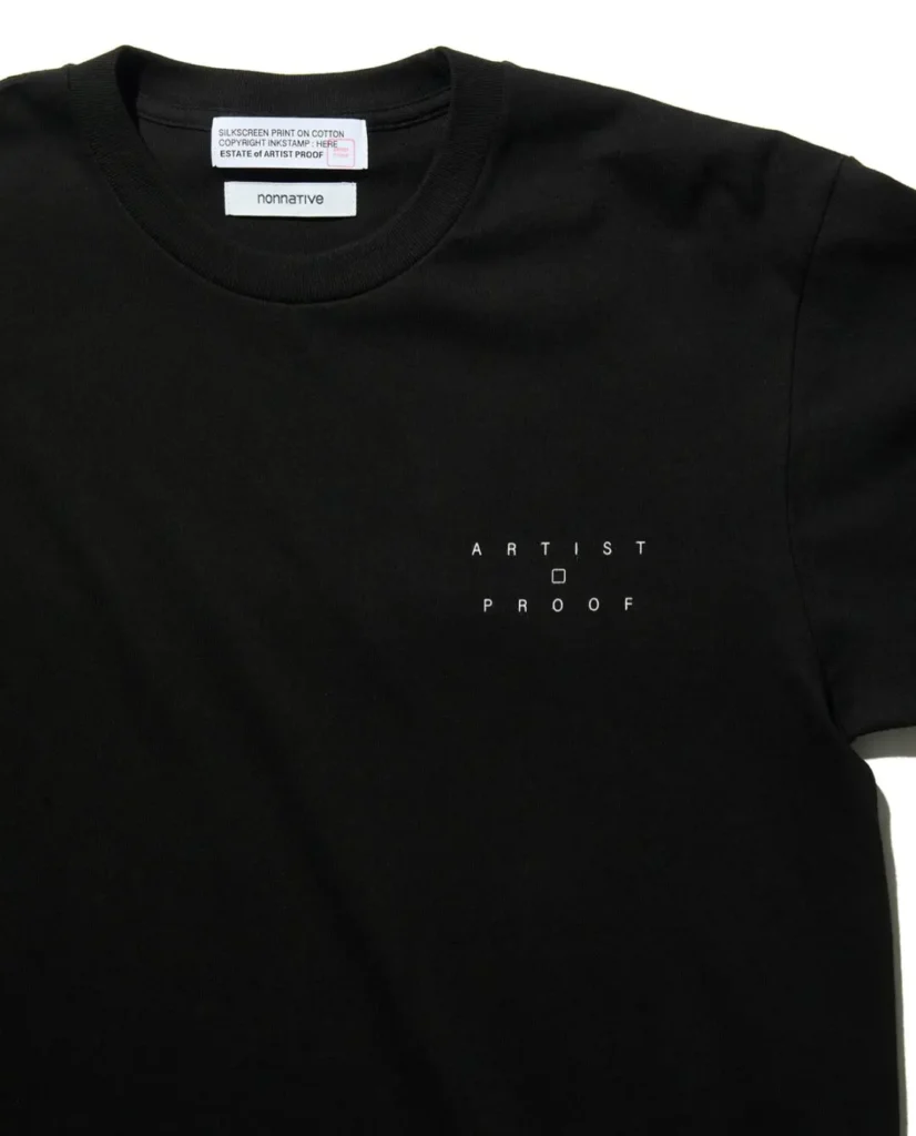 Close-up of the black T-shirt chest showing the small "ARTIST PROOF" logo, nonnative neck label, and printed collide tag inside the collar