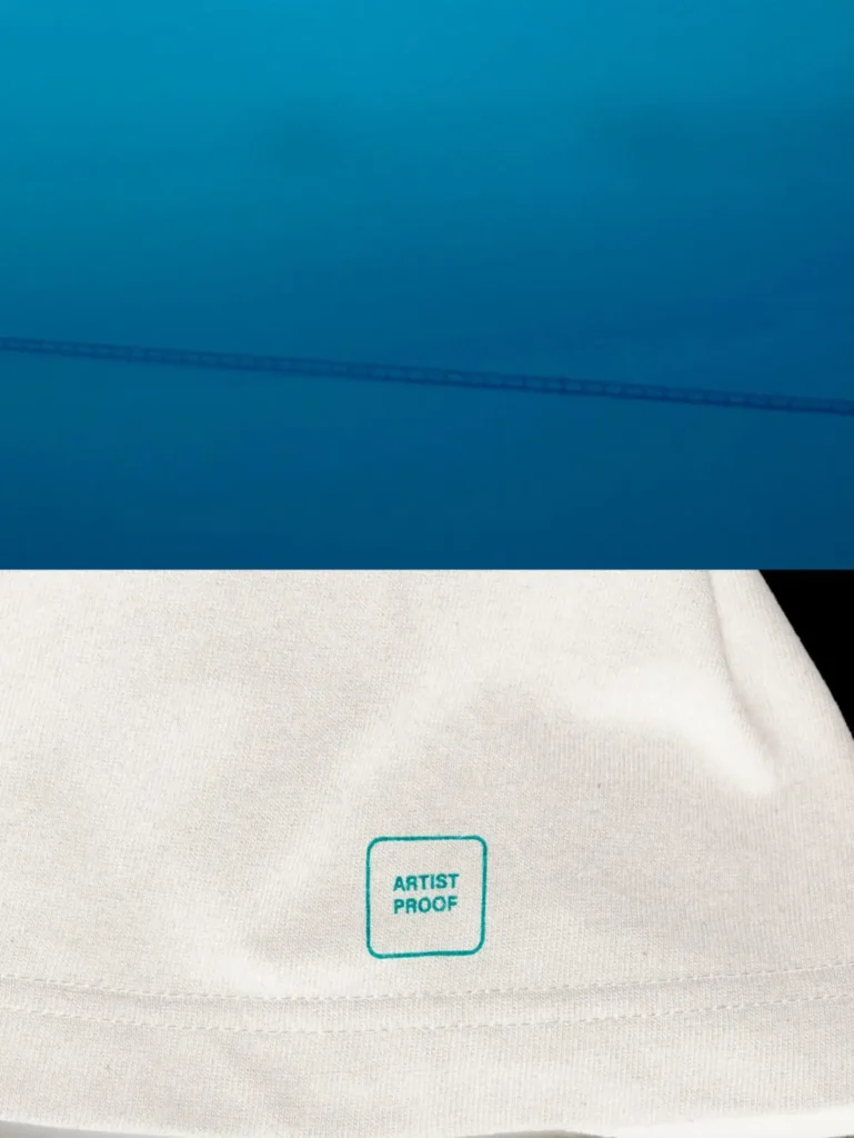Blue-toned abstract artwork featuring a faint linear structure above a close-up of the white T-shirt hem with a turquoise square "ARTIST PROOF" stamp