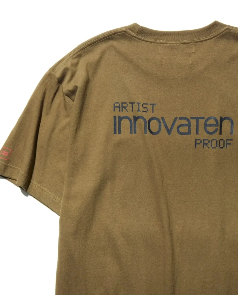 Olive green T-shirt back featuring large "ARTIST innovaten PROOF" typography in navy, with subtle collide graphics on the sleeve