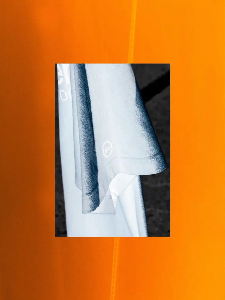 White T-shirts hanging on a rack with a Fragment Design lightning bolt sleeve logo, framed against a vivid orange abstract backdrop