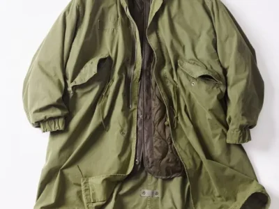 Front view of an olive green fishtail parka displayed open with its removable olive quilted liner attached, featuring oversized flap pockets, a stand collar, front zipper, snap closure, and adjustable fishtail hem