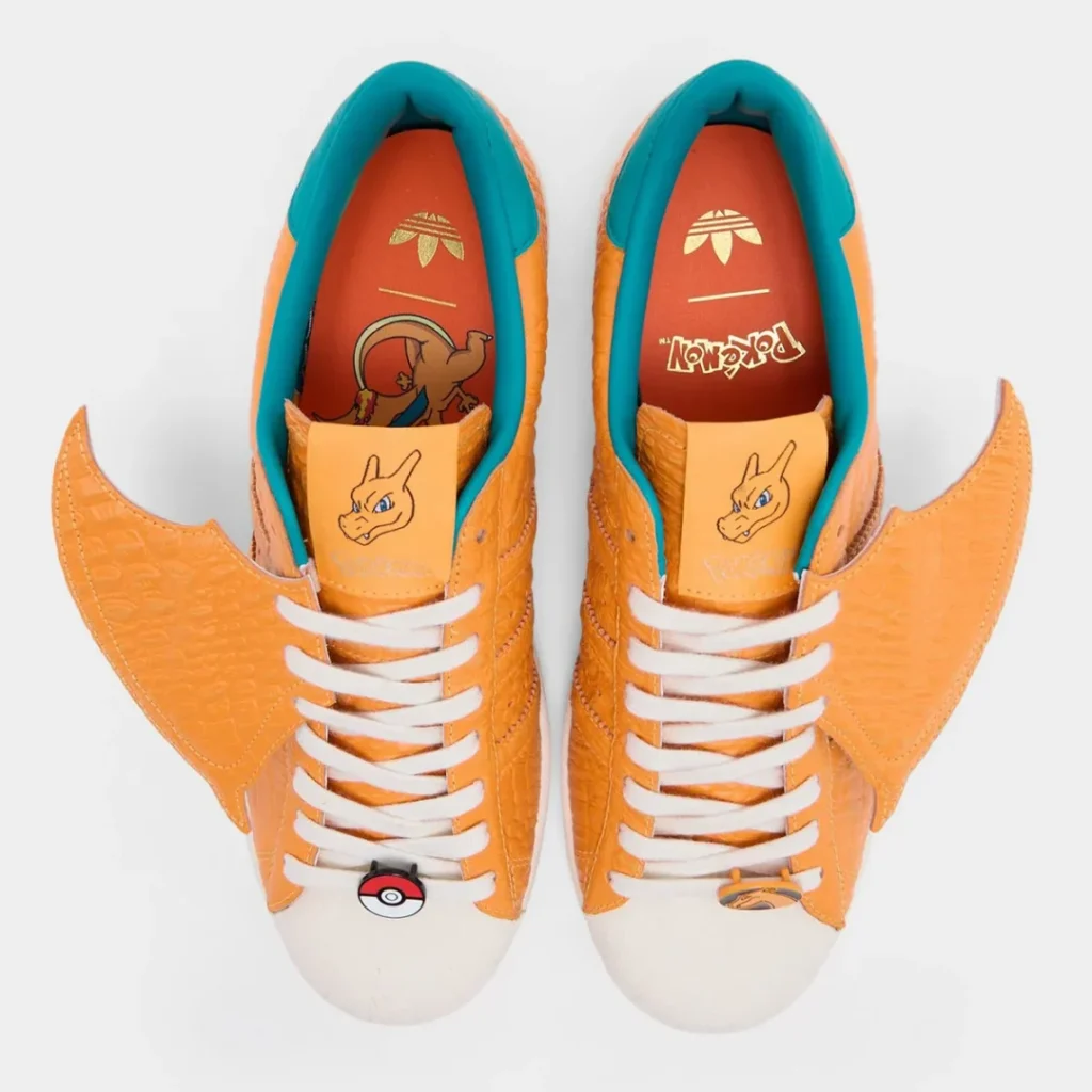 Top-down view of the Charizard-inspired adidas shoe showcasing illustrated tongue logos, teal interiors, Poké Ball lace jewel, and oversized wing detail