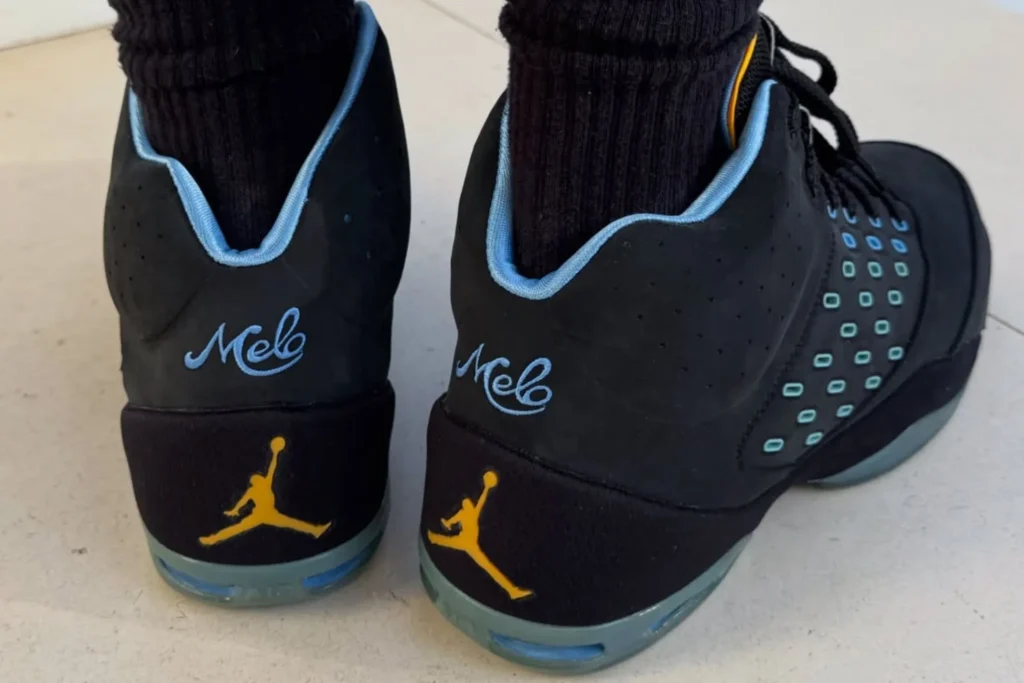 Rear view of a pair of black Jordan Melo basketball shoes worn with black crew socks. The heels feature powder blue "Melo" script branding above gold Jumpman logos, while translucent blue-green Air cushioning is visible in the heel. Light blue collar lining and subtle yellow accents complete the design