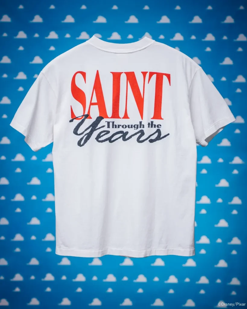White Tee displayed from the back with oversized red and black "Saint Through the Years" graphic on a blue Toy Story cloud backdrop