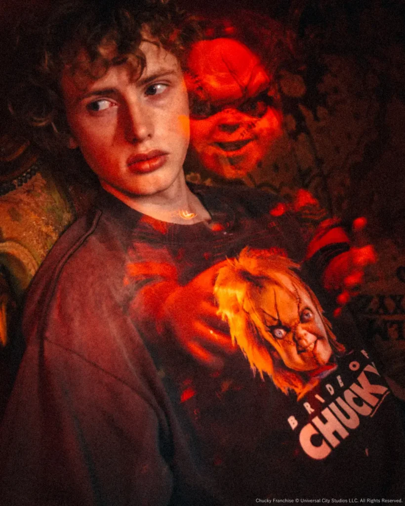 Close-up editorial portrait of a model in the "Bride of Chucky" Tee with a red-lit Chucky doll positioned directly behind, creating a suspenseful horror atmosphere