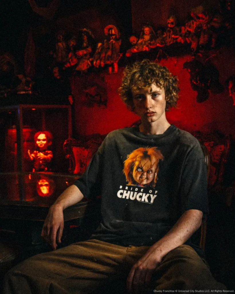 Wide editorial shot of a model seated in a vintage chair wearing the oversized "Bride of Chucky" tee, surrounded by shelves of collectible horror dolls in dim red light