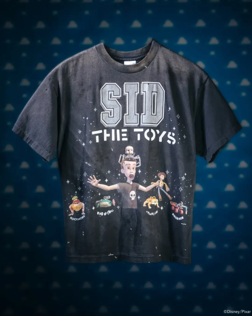 Flat-lay style front product image of the faded charcoal "Sid the Toys" T-shirt featuring Sid Phillips and iconic Toy Story toys on a blue cloud-pattern backdrop
