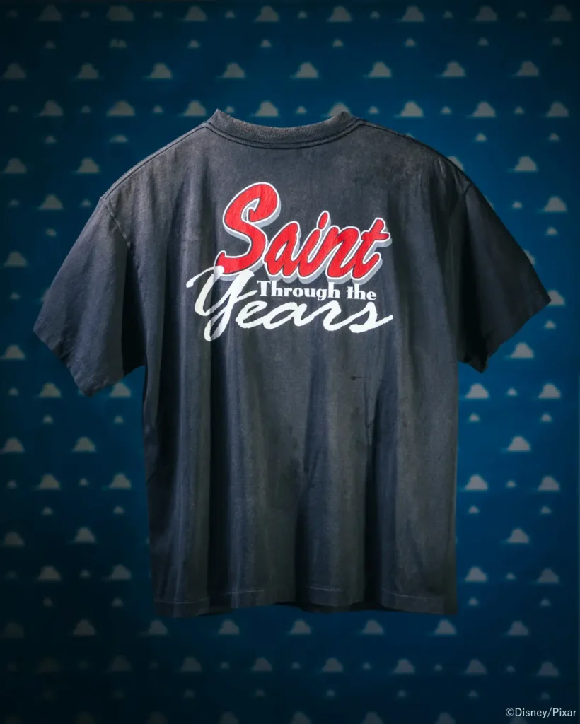 Back product image of the faded charcoal "Saint Through the Years" Tee featuring oversized red, white, and gray script graphics against a blue Toy Story cloud backdrop