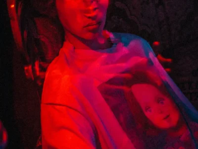 Model reclining in an oversized white graphic Tee featuring a vintage-style Charles Lee Ray portrait, while a red-lit Chucky doll looms behind in a dark, horror-inspired room