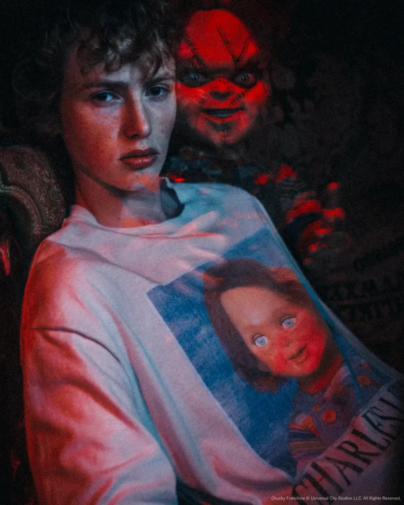 Close-up of a seated model wearing the oversized Charles Lee Ray graphic tee under dramatic red and blue lighting, with Chucky peering over their shoulder for a cin horror effect