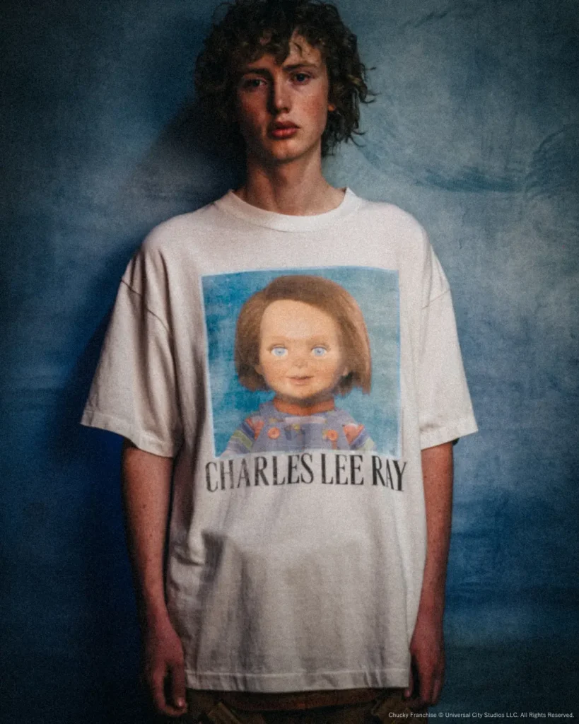 Front view of a model wearing an oversized white Charles Lee Ray T-shirt against a textured blue backdrop, showcasing the large vintage mugshot-style Chucky graphic