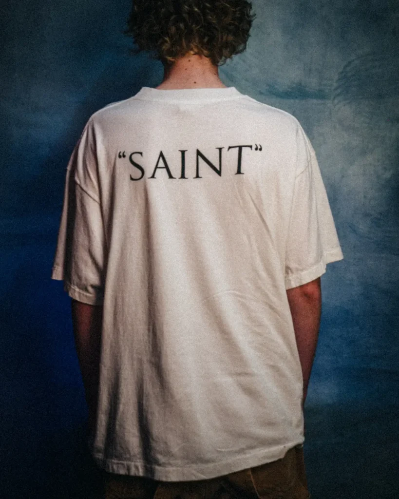 Rear view of the oversized white Tee displaying bold black “SAINT” lettering across the upper back against a muted blue studio background