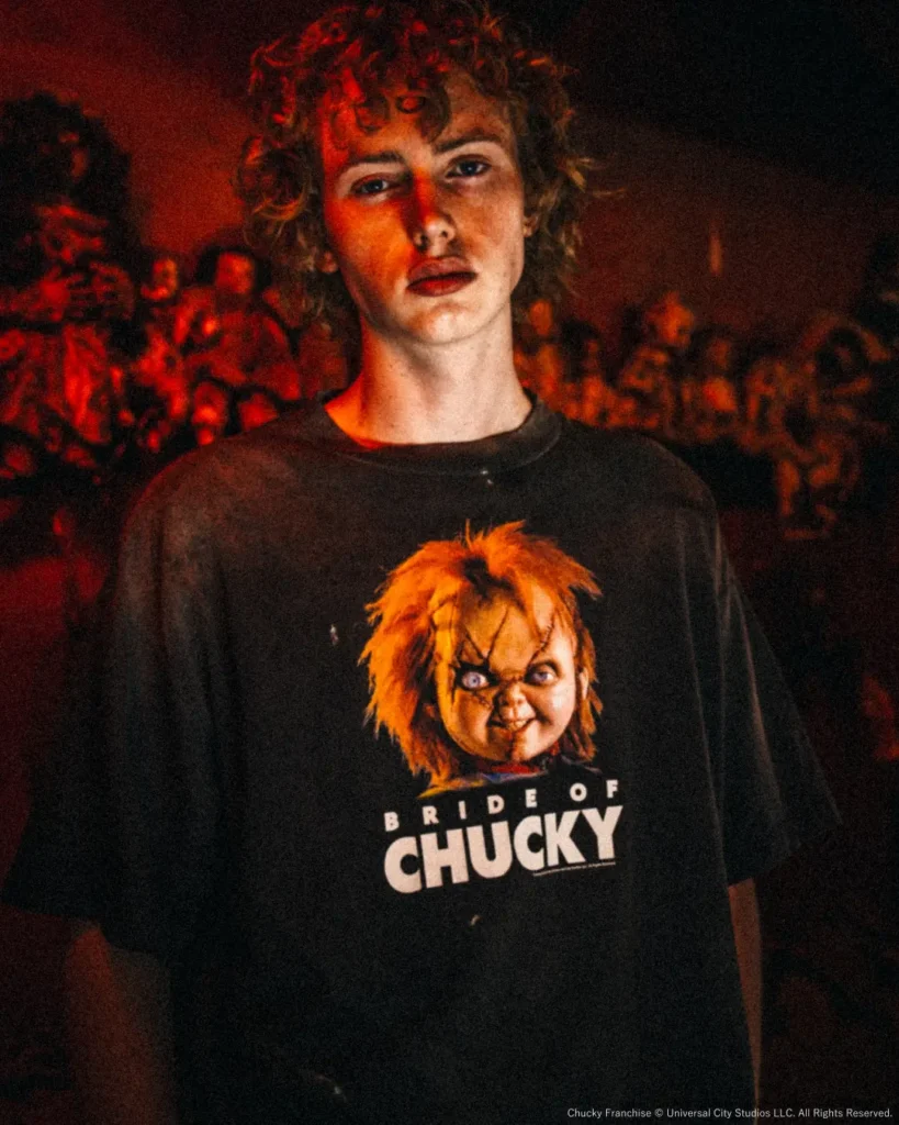 Portrait of a model wearing a faded black oversized "Bride of Chucky" graphic T-shirt in a dim room filled with horror dolls and warm red ambient light