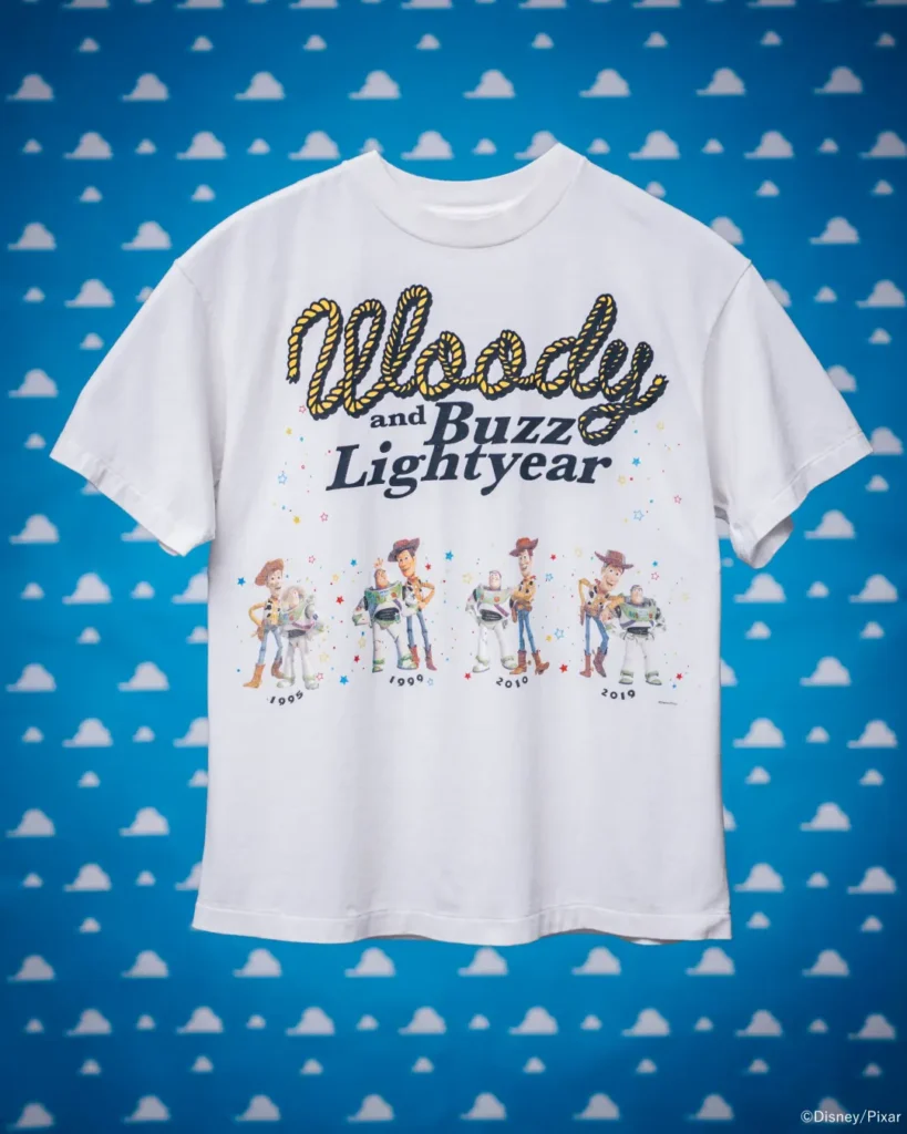 White Toy Story-inspired Tee displayed flat against a blue cloud-pattern backdrop, featuring "Woody and Buzz Lightyear" typography with character artwork spanning multiple film era