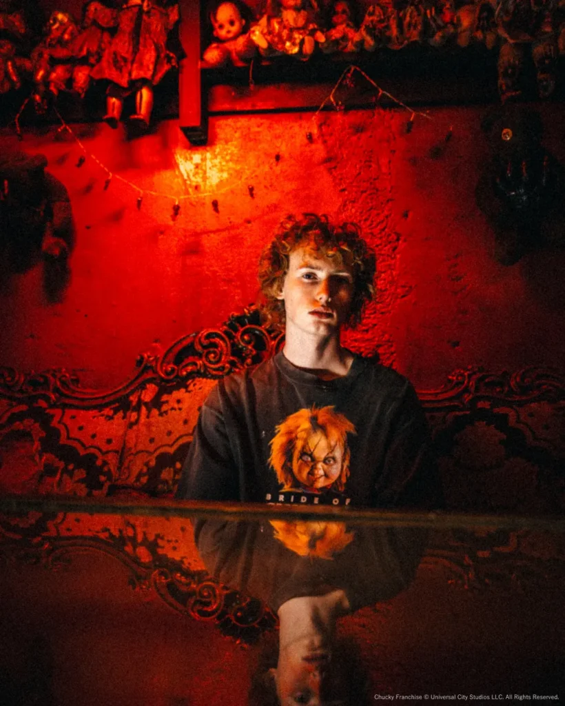 Model seated in a haunted doll-filled room wearing the "Bride of Chucky" tee, reflected across a glossy tabletop under moody red light