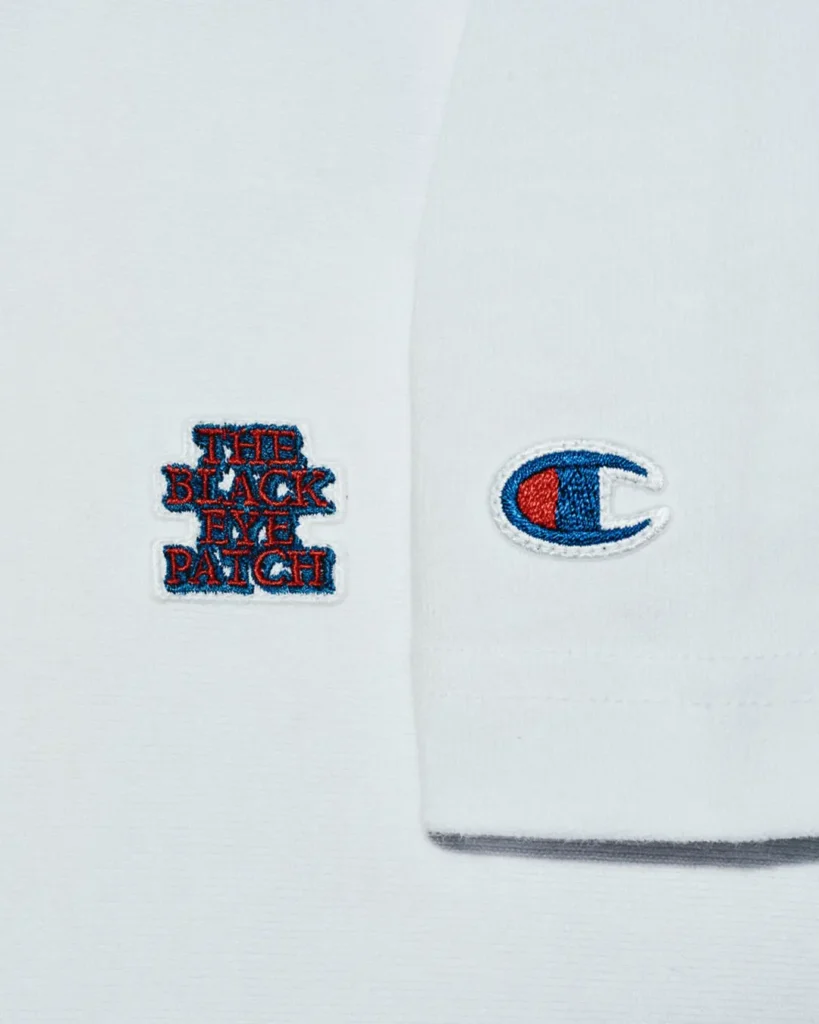 Close up of the white Tee featuring an embroidered The Black Eye Patch logo on the chest with the classic Champion C logo embroidered on the sleeve