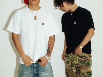 Two models wearing the collision Tee, one in white with light distressed denim and a red cap, the other in black with camouflage cargo pants, highlighting the minimalist embroidered chest logo