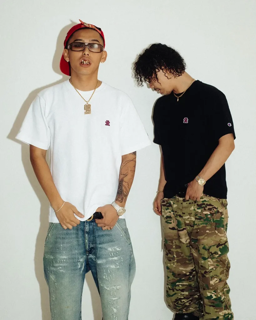 Two models wearing the collision Tee, one in white with light distressed denim and a red cap, the other in black with camouflage cargo pants, highlighting the minimalist embroidered chest logo