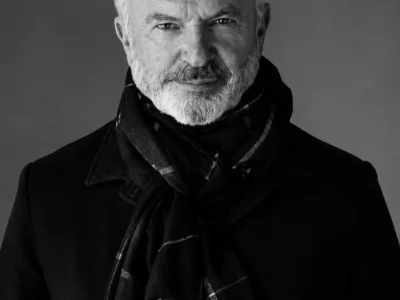 A high-resolution studio portrait of Sam Neill with silver hair and a neatly trimmed white beard, wearing a dark overcoat and navy check-pattern scarf against a clean white background. He faces the camera with a calm, confident expression under soft, even lighting, creating a polished editorial-style headshot